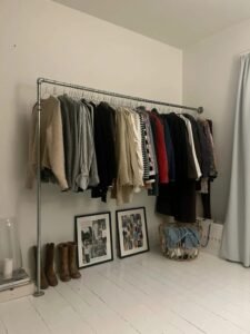 Freestanding industrial galvanised pipe clothing rack holding a row of jackets and knitwear against a white wall